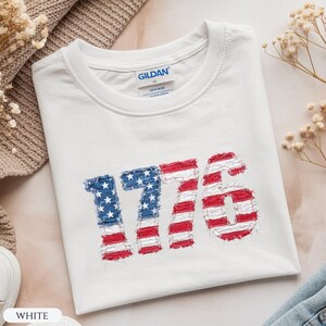 May include: White t-shirt with the number "1776" in a distressed American flag design. The "1" and "7" are blue with white stars, and the "7" and "6" are red and white stripes. The shirt has a crew neck.