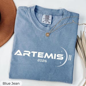 May include: A blue jean colored t-shirt with the word "ARTEMIS" and the year "2026" printed in white. A gold necklace with a circular pendant is draped across the shirt. The shirt is folded on a white surface.