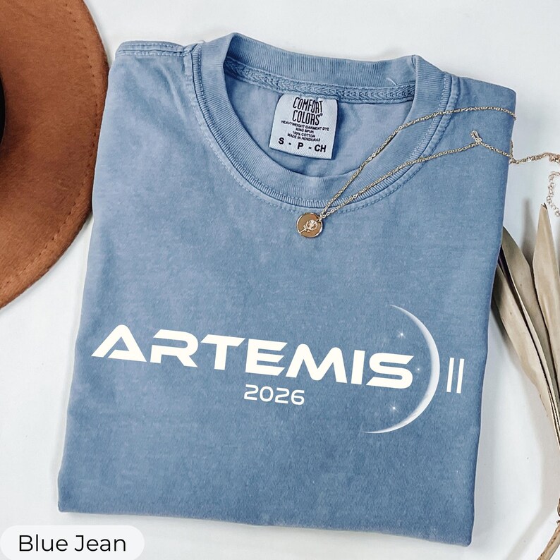 May include: A blue jean colored t-shirt with the word "ARTEMIS" and the year "2026" printed in white. A gold necklace with a circular pendant is draped across the shirt. The shirt is folded on a white surface.
