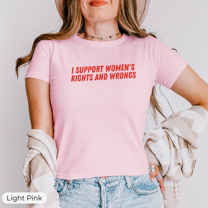 May include: Light pink t-shirt with the text "I SUPPORT WOMEN'S RIGHTS AND WRONGS" in red. The shirt is short-sleeved and made of a soft material. The model is wearing blue jeans and a beige hat.