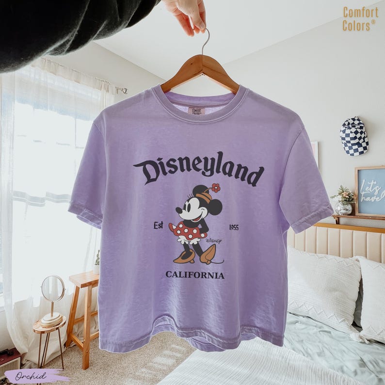 Puede incluir: Una camiseta Comfort Colors lavanda con un gr&aacute;fico de Disneyland con Minnie Mouse. El dise&ntilde;o incluye el texto "Disneyland", "Est 1955" y "California". Minnie Mouse se representa con una falda roja de lunares y zapatos marrones.