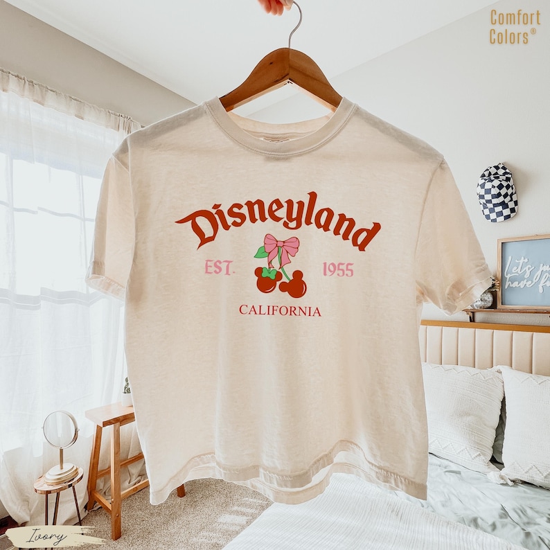 May include: Ivory-colored t-shirt with the word "Disneyland" in a red script font. Below the word is "EST. 1955" and "CALIFORNIA" in a pink font. A graphic of cherries with a pink bow is in the center.