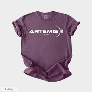 May include: Berry-colored t-shirt with the word "ARTEMIS" in white, along with the year "2026" and a crescent moon design. The shirt has short sleeves and a crew neck.