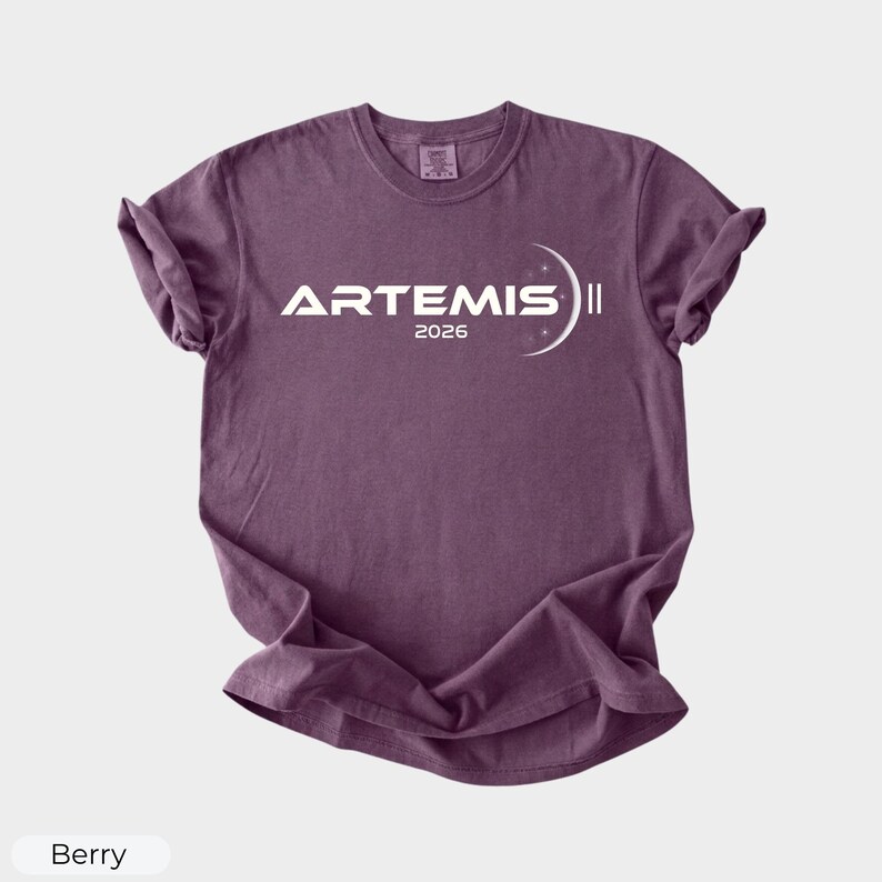 May include: Berry-colored t-shirt with the word "ARTEMIS" in white, along with the year "2026" and a crescent moon design. The shirt has short sleeves and a crew neck.