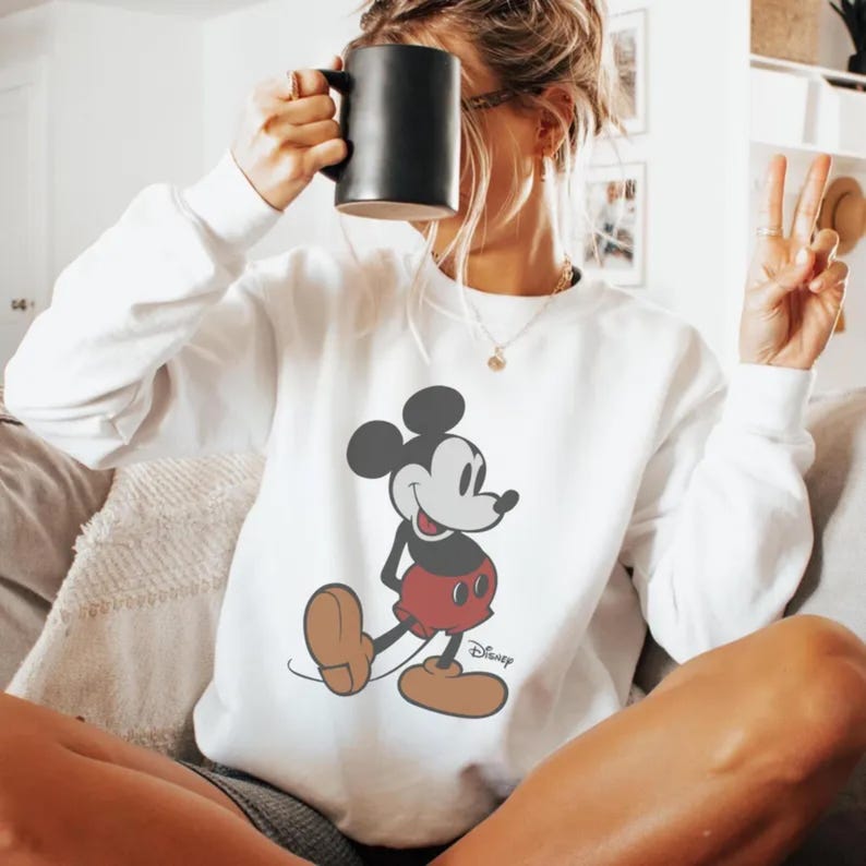 Puede incluir: Sudadera blanca con un gr&aacute;fico de Mickey Mouse. El personaje de dibujos animados est&aacute; en negro, rojo y marr&oacute;n. El logotipo de Disney est&aacute; impreso debajo. La sudadera es de manga larga y tiene cuello redondo. Se sostiene una taza negra.