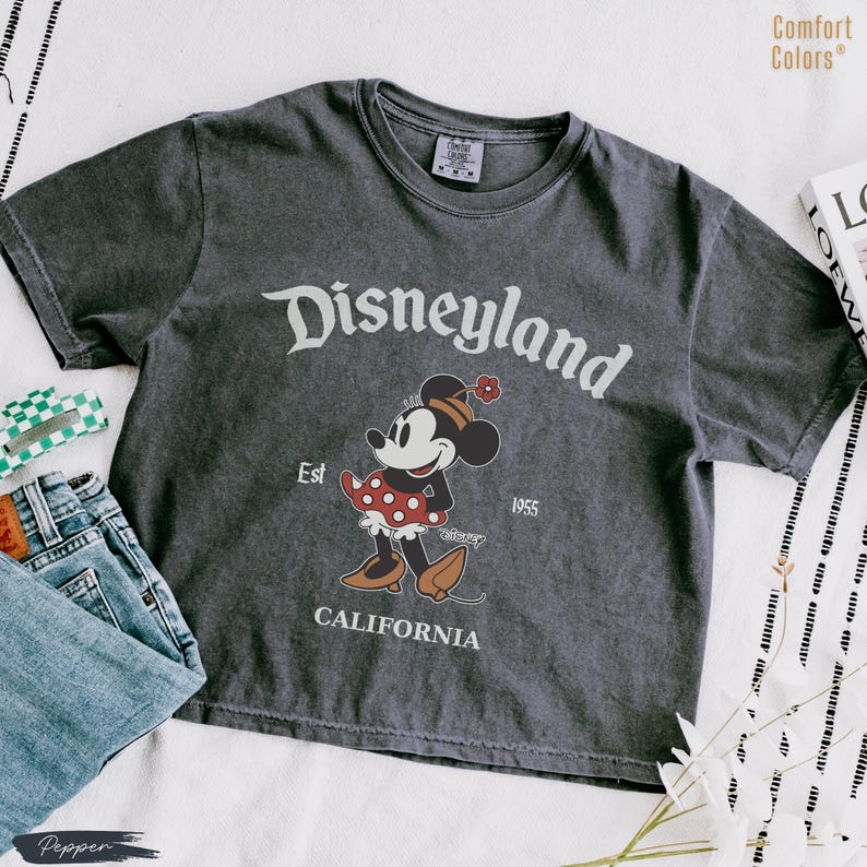 Puede incluir: Camiseta gris oscuro con la palabra "Disneyland" en escritura blanca. La camiseta presenta un gr&aacute;fico de Minnie Mouse y el texto "CALIFORNIA" y "Est 1955". La camiseta se combina con unos vaqueros azules y una pinza para el pelo a cuadros.