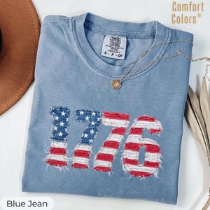 May include: Blue jean colored t-shirt with the number "1776" in a distressed American flag design. The shirt has a crew neck and short sleeves. A gold necklace with a pendant is draped across the shirt.