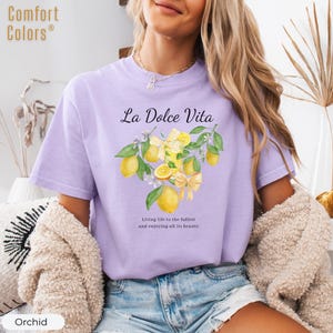 K&ouml;nnte beinhalten: Orchideenfarbenes T-Shirt mit dem Text "La Dolce Vita" und einer Aquarellillustration von Zitronen und Schleifen. Das Shirt tr&auml;gt auch den Text "Das Leben in vollen Z&uuml;gen genie&szlig;en und all seine Sch&ouml;nheit genie&szlig;en."