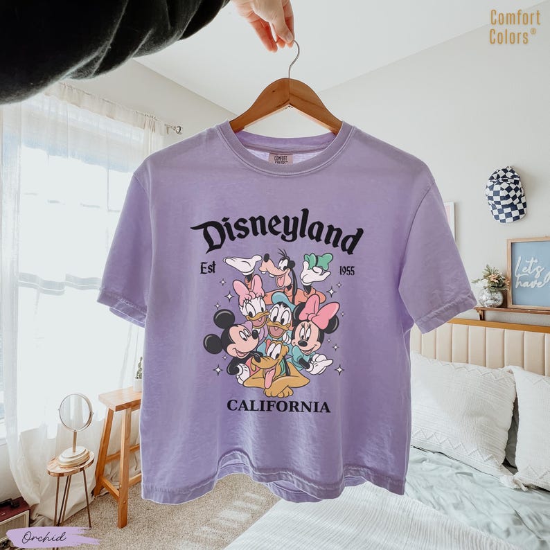 May include: A lavender Comfort Colors t-shirt featuring a Disneyland graphic. The graphic includes Mickey Mouse, Minnie Mouse, Donald Duck, Daisy Duck, Goofy, and Pluto. The text "Disneyland Est 1955 California" is printed above and below the characters.