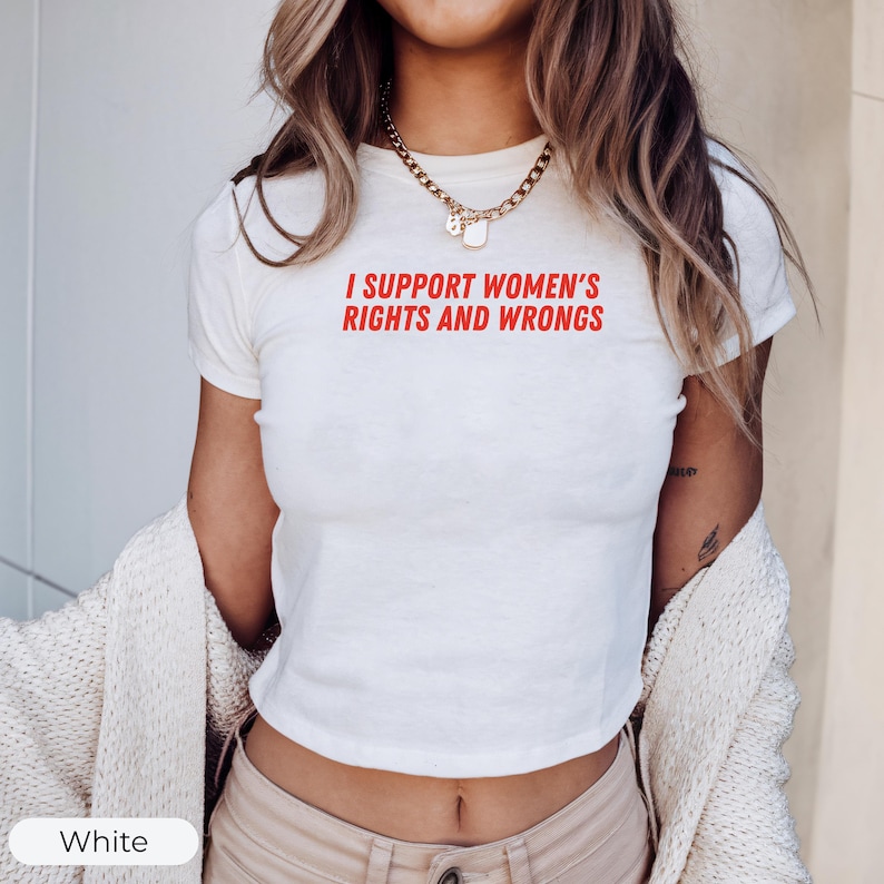May include: White crop top with the red text "I SUPPORT WOMEN'S RIGHTS AND WRONGS". The shirt has short sleeves and a crew neck. A gold necklace is worn around the neck. A white cardigan is draped over the shoulders.