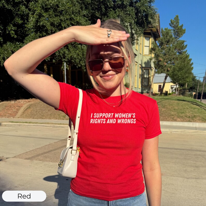 May include: A red t-shirt with the text "I SUPPORT WOMEN'S RIGHTS AND WRONGS" in white. The shirt is short-sleeved and worn by a person wearing sunglasses and a small white purse. The background includes a house and trees.