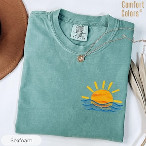 Puede incluir: Camiseta verde mar con un gráfico de un sol amarillo que se pone sobre olas azules. La camiseta tiene cuello redondo y mangas cortas. Un collar plateado con un pequeño colgante está drapeado sobre la camiseta. La etiqueta dice "Comfort Colors".