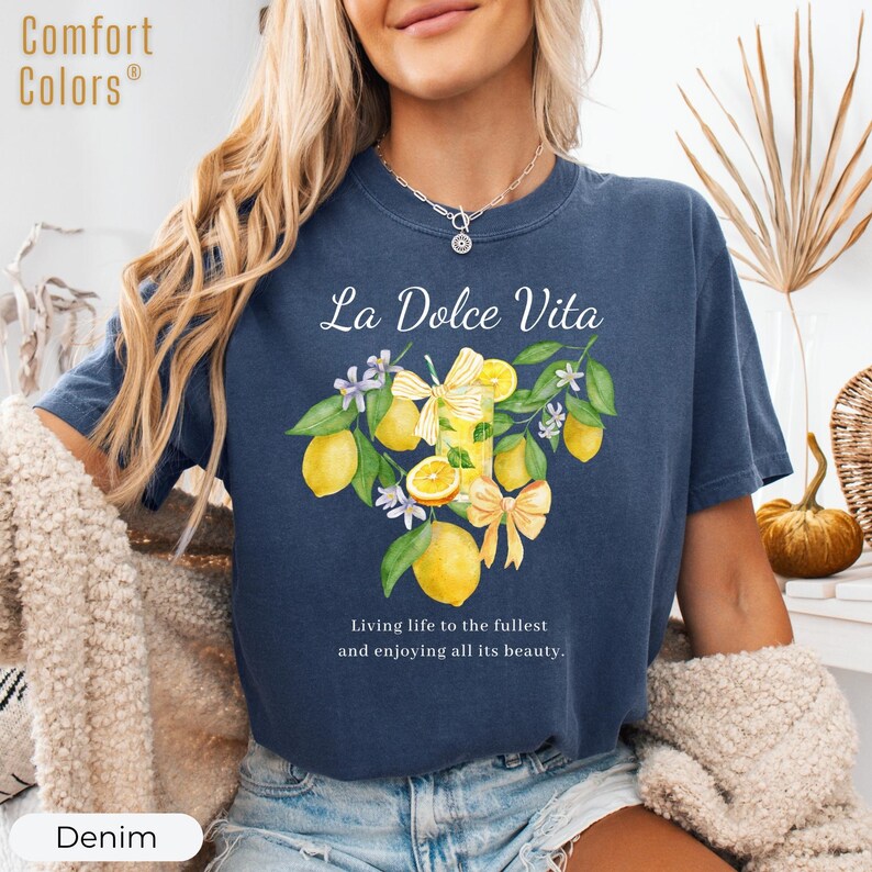 K&ouml;nnte beinhalten: Denimblaues T-Shirt mit dem Text "La Dolce Vita" und einer Aquarellillustration von Zitronen, Blumen und einem Getr&auml;nk. Das Shirt enth&auml;lt auch den Text "Living life to the fullest and enjoying all its beauty."