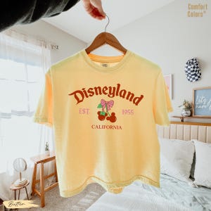 May include: A yellow t-shirt with the word "Disneyland" in a cursive font, along with the text "EST. 1955 CALIFORNIA". The graphic includes a pink bow and cherries. The shirt is hanging on a wooden hanger.