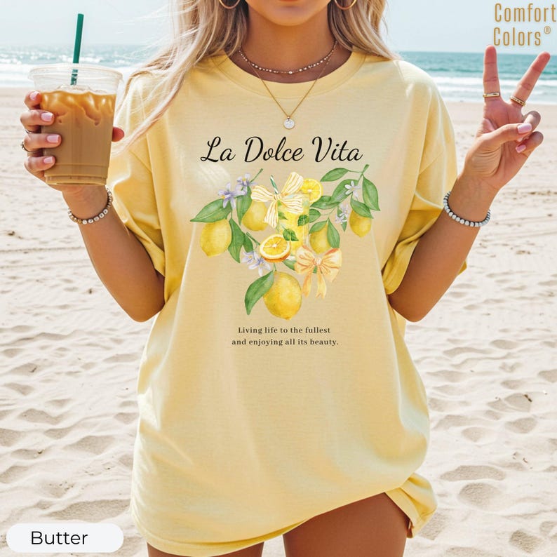 K&ouml;nnte beinhalten: Hellgelbes T-Shirt mit dem Schriftzug "La Dolce Vita" und einem Aquarell-Zitronen-Design. Der Text "Das Leben in vollen Z&uuml;gen genie&szlig;en und all seine Sch&ouml;nheit genie&szlig;en" ist ebenfalls vorhanden. Die Marke Comfort Colors ist sichtbar.