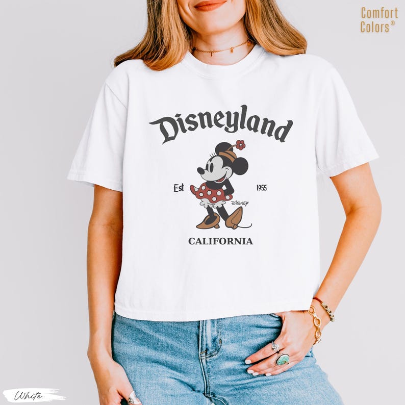 Puede incluir: Camiseta blanca con un dise&ntilde;o vintage de Disneyland. El dise&ntilde;o incluye el logotipo de Disneyland, Minnie Mouse, y el texto "CALIFORNIA" y "Est 1955". La camiseta es de la marca Comfort Colors.