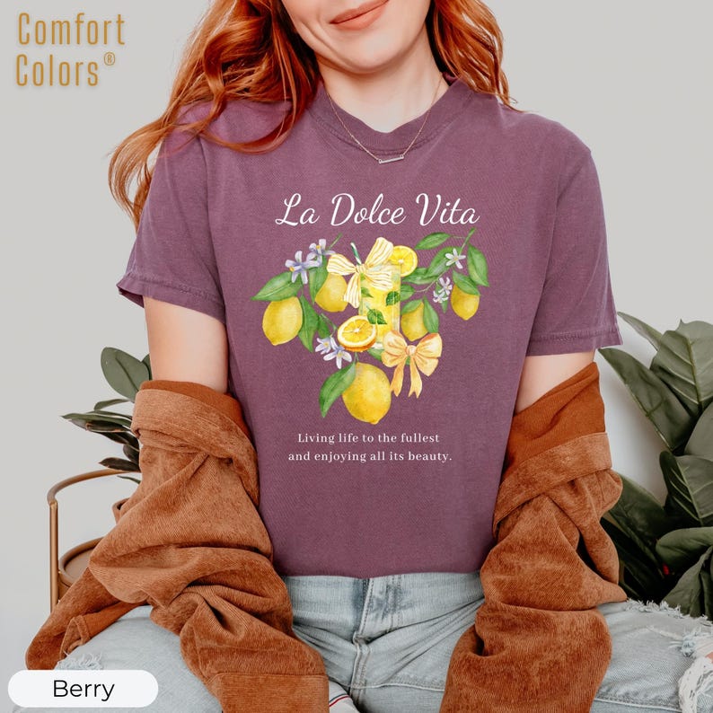 K&ouml;nnte beinhalten: Beerenfarbenes T-Shirt mit dem Text "La Dolce Vita" und einer Aquarellillustration von Zitronen, Blumen und Schleifen. Das Shirt hat auch den Text "Das Leben in vollen Z&uuml;gen genie&szlig;en und all seine Sch&ouml;nheit genie&szlig;en."