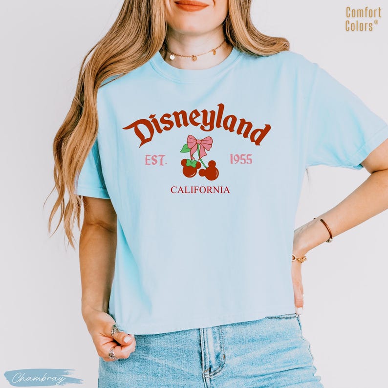 May include: Light blue t-shirt with the word "Disneyland" in a cursive font, along with "EST. 1955 CALIFORNIA" in a serif font. A graphic of cherries with a pink bow is centered below the word "Disneyland".