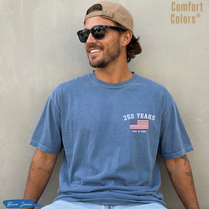 May include: A blue short-sleeve t-shirt featuring a graphic on the chest with the text 250 YEARS above a United States flag and the text 1776 2026 below it. The shirt has a crew neckline and a solid, muted blue color throughout the fabric.
