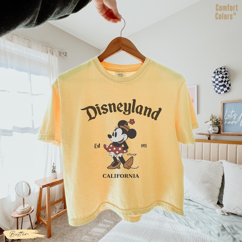 Puede incluir: Camiseta amarilla mantequilla con el logotipo de Disneyland y un gr&aacute;fico de Minnie Mouse. El gr&aacute;fico incluye el texto "Est 1955" y "California". La camiseta est&aacute; colgada en una percha de madera.