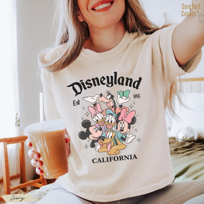 May include: Ivory-colored t-shirt featuring a Disneyland graphic with Mickey Mouse, Minnie Mouse, Donald Duck, Daisy Duck, Goofy, and Pluto. The text "Disneyland Est 1955 California" is printed above and below the characters. The shirt is a Comfort Colors brand.