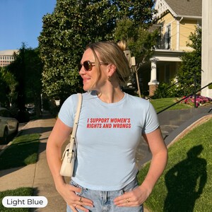 May include: Light blue t-shirt with the text "I SUPPORT WOMEN'S RIGHTS AND WRONGS" in red. The shirt is paired with blue jeans. The image is taken outdoors on a sunny day.