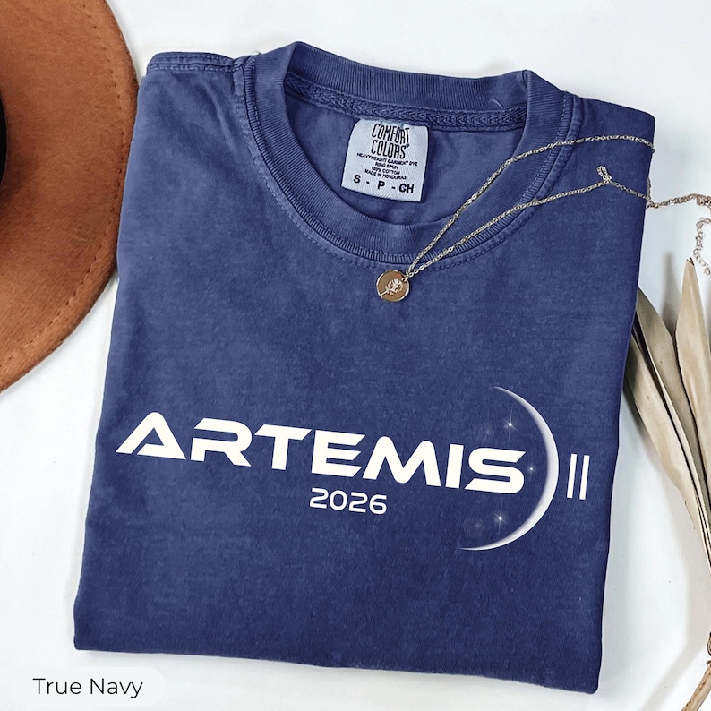 May include: A navy blue t-shirt with the word "ARTEMIS II" and the year "2026" printed in white. A gold necklace with a pendant is draped across the shirt. The shirt is folded on a white surface.