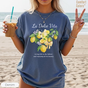 K&ouml;nnte beinhalten: Denimblaues T-Shirt mit dem Text "La Dolce Vita" und einem Zitronen- und Blumendesign. Das Shirt hat auch den Text "Living life to the fullest and enjoying all its beauty." Das Shirt ist von der Marke Comfort Colors.