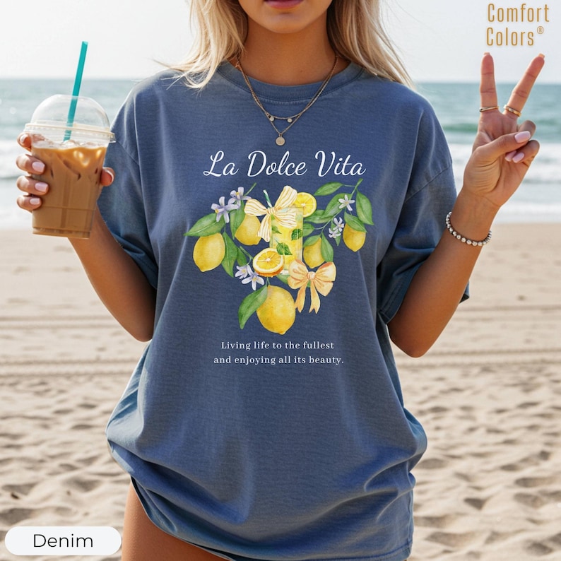 K&ouml;nnte beinhalten: Denimblaues T-Shirt mit dem Text "La Dolce Vita" und einem Zitronen- und Blumendesign. Das Shirt hat auch den Text "Living life to the fullest and enjoying all its beauty." Das Shirt ist von der Marke Comfort Colors.