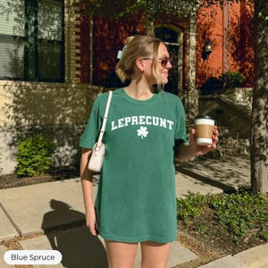 May include: A green t-shirt with the text "LEPRECONT" in white, and a white shamrock graphic. The shirt is a solid color and has short sleeves. The person is holding a coffee cup and a white purse.