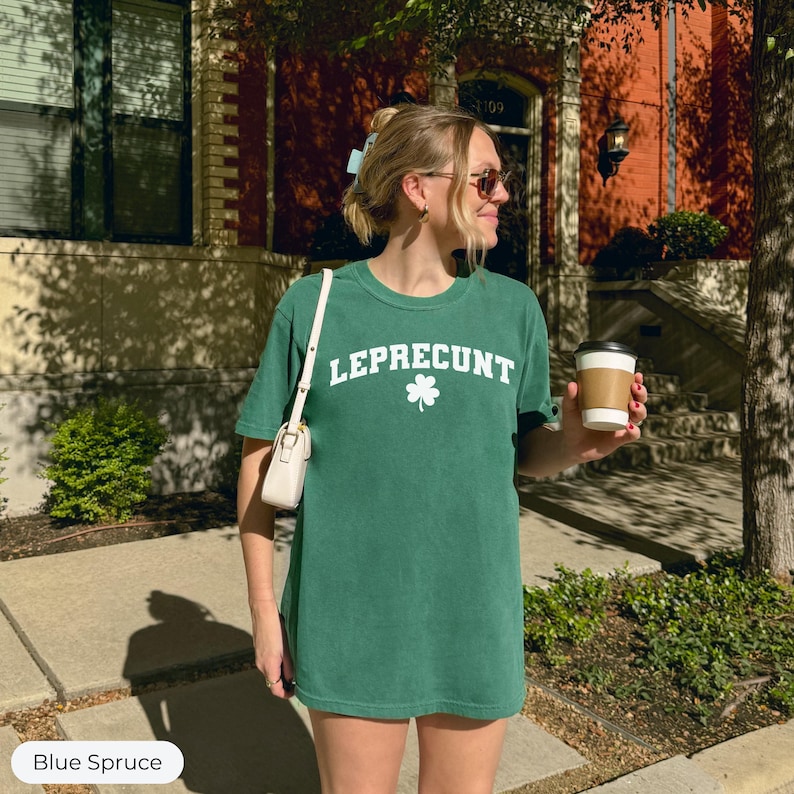 May include: A green t-shirt with the text "LEPRECONT" in white, and a white shamrock graphic. The shirt is a solid color and has short sleeves. The person is holding a coffee cup and a white purse.
