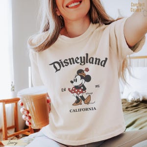 Puede incluir: Camiseta de color marfil con un gr&aacute;fico de Disneyland. El dise&ntilde;o incluye la palabra "Disneyland" en una fuente de escritura, el texto "Est 1955" y un personaje de dibujos animados de Minnie Mouse. La palabra "CALIFORNIA" tambi&eacute;n est&aacute; impresa en la camiseta.