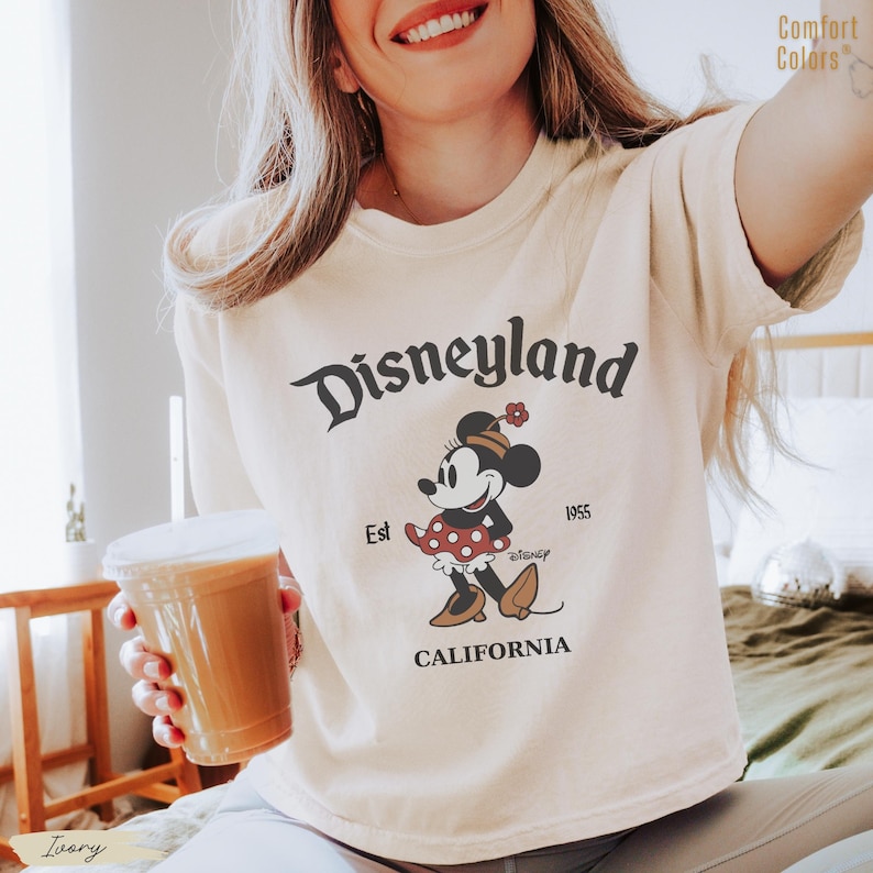 Puede incluir: Camiseta de color marfil con un gr&aacute;fico de Disneyland. El dise&ntilde;o incluye la palabra "Disneyland" en una fuente de escritura, el texto "Est 1955" y un personaje de dibujos animados de Minnie Mouse. La palabra "CALIFORNIA" tambi&eacute;n est&aacute; impresa en la camiseta.