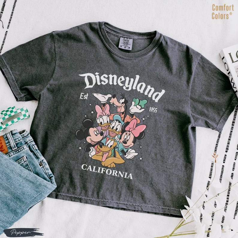 May include: Dark gray t-shirt with a Disneyland graphic featuring Mickey Mouse, Minnie Mouse, Donald Duck, Daisy Duck, Goofy, and Pluto. The text "Disneyland Est 1955 California" is printed in white. The shirt is paired with blue jeans.
