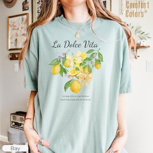 K&ouml;nnte beinhalten: Hellblaues Comfort Colors T-Shirt mit dem Text "La Dolce Vita" &uuml;ber einer Aquarellillustration von Zitronen, Schleifen und Bl&auml;ttern. Der Text darunter lautet: "Das Leben in vollen Z&uuml;gen genie&szlig;en und all seine Sch&ouml;nheit genie&szlig;en."