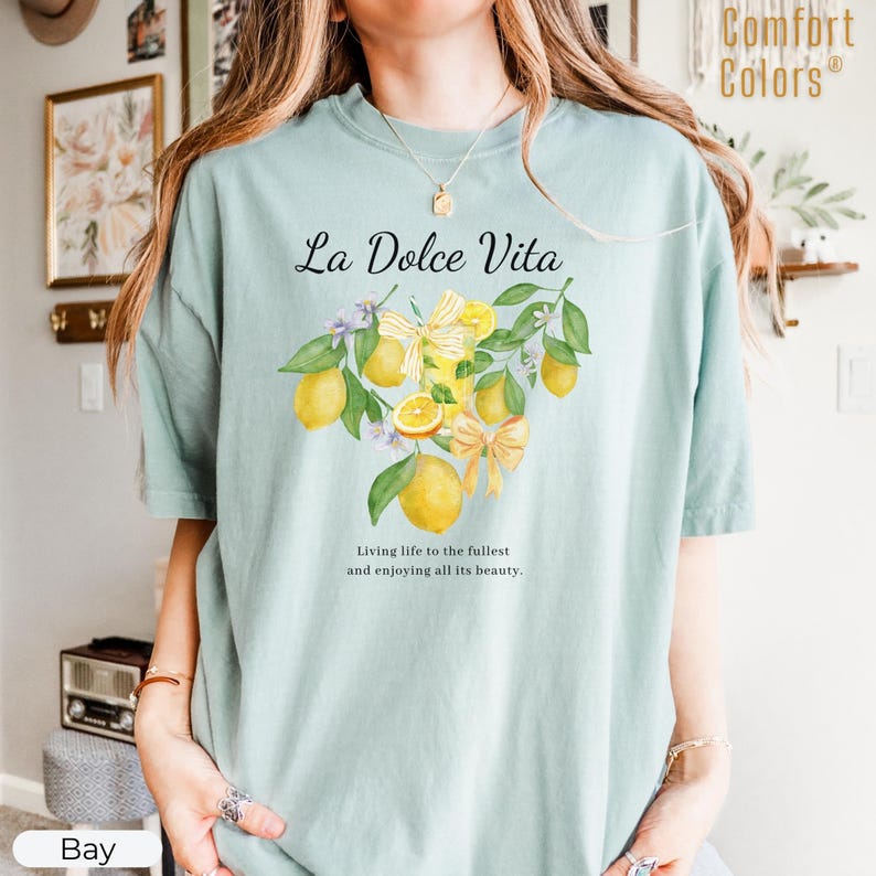 K&ouml;nnte beinhalten: Hellblaues Comfort Colors T-Shirt mit dem Text "La Dolce Vita" &uuml;ber einer Aquarellillustration von Zitronen, Schleifen und Bl&auml;ttern. Der Text darunter lautet: "Das Leben in vollen Z&uuml;gen genie&szlig;en und all seine Sch&ouml;nheit genie&szlig;en."