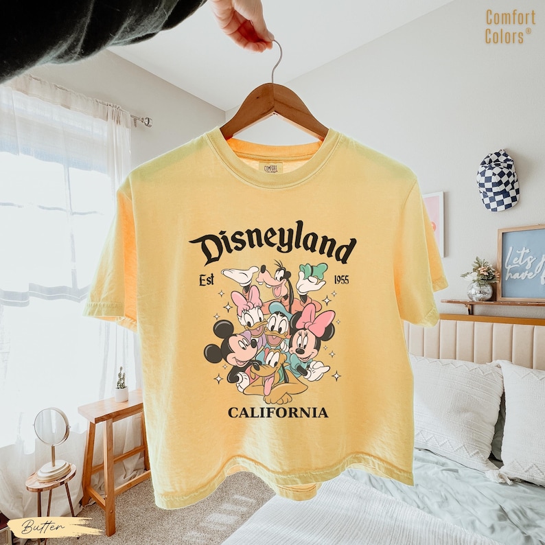 May include: Yellow short-sleeved t-shirt with a Disneyland graphic featuring Mickey Mouse, Minnie Mouse, Donald Duck, Daisy Duck, Goofy, and Pluto. The text "Disneyland Est 1955 California" is printed in black.