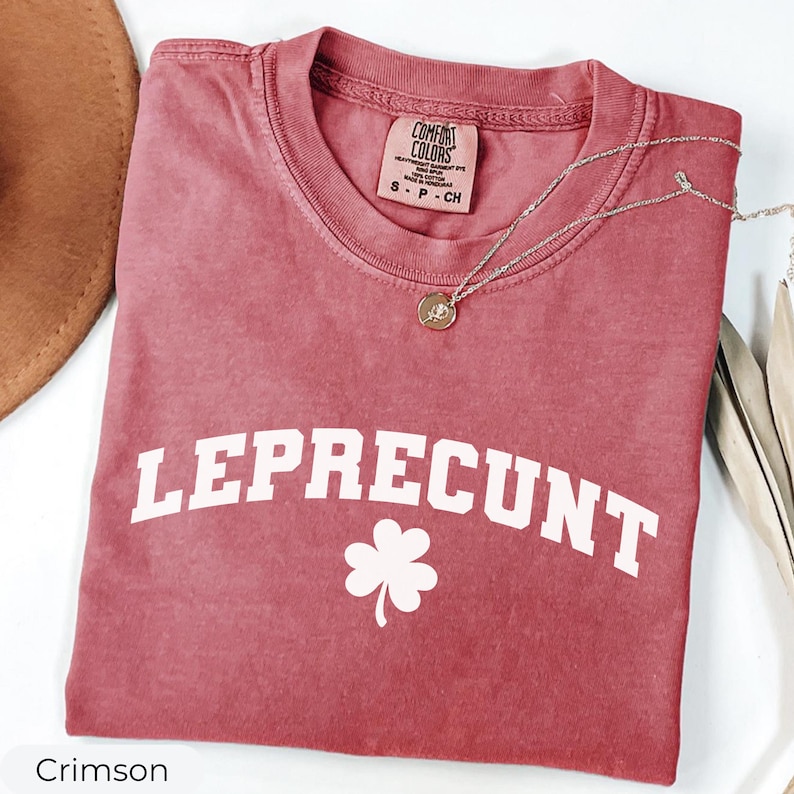 May include: Crimson-colored t-shirt with the word "LEPRECUNT" in white block letters and a white shamrock. A silver necklace with a small pendant rests on the shirt. The shirt has a Comfort Colors tag.