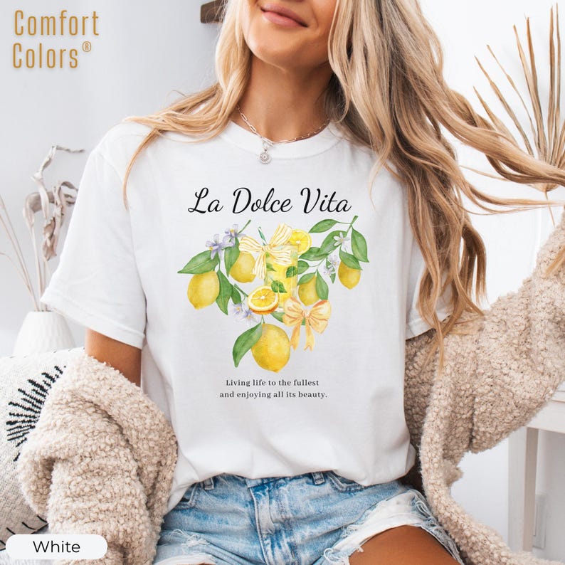 K&ouml;nnte beinhalten: Wei&szlig;es T-Shirt mit dem Text "La Dolce Vita" und einer Aquarellillustration von Zitronen, Bl&auml;ttern und Schleifen. Das Shirt hat auch den Text "Das Leben in vollen Z&uuml;gen genie&szlig;en und all seine Sch&ouml;nheit genie&szlig;en."