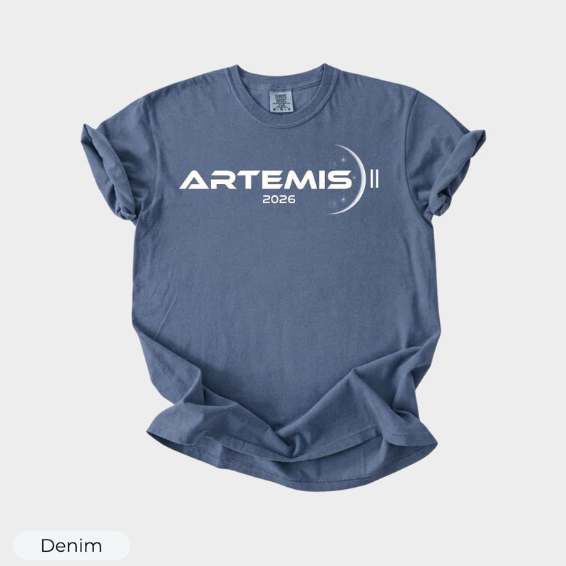 May include: Denim blue t-shirt with the word "ARTEMIS" in white, along with the year "2026" and a crescent moon graphic. The shirt is a crew neck and has rolled sleeves.