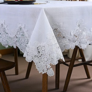 Op de afbeelding: Een wit tafelkleed met ingewikkelde bloemenkanten rand. Het kantontwerp heeft gedetailleerde bloemen en bladeren, wat een delicate touch toevoegt aan de tafelsetting. Het tafelkleed is gedrapeerd over een houten tafel, wat een elegante en verfijnde uitstraling creëert.