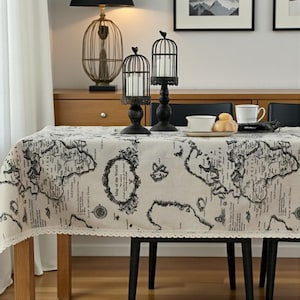 May include: A cream-colored tablecloth with a black world map design and a lace trim. Two black metal candle holders with birdcage designs and candles are on the table. A lamp and other decorative items are in the background.