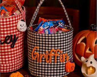 Puede incluir: Un cubo de dulces de Halloween de tela de cuadros blancos y negros con el nombre "Griffin" en letras naranjas. El cubo está lleno de caramelos y tiene un asa. Una calabaza naranja tallada y un portavelas fantasma están cerca.