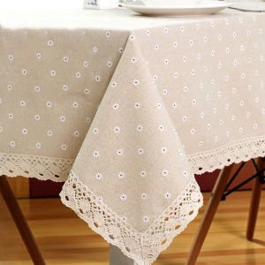 May include: Beige tablecloth with a repeating pattern of small white daisies. The edges are trimmed with a delicate, cream-colored lace. The tablecloth is draped over a wooden table, creating a rustic and charming aesthetic.