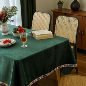 Puede incluir: Una mesa de comedor con un mantel verde oscuro y dos sillas con respaldos de mimbre. La mesa está puesta con un vaso de líquido ámbar, un cuenco de fruta y un plato de tomates. Un jarrón con flores y un libro abierto también están presentes.
