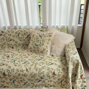 May include: A sofa with a cream-coloured bird and floral patterned cover and matching throw pillows. The throw pillows include a square pillow with a bird and floral pattern and a fluffy white pillow. The sofa is near a window with white curtains.