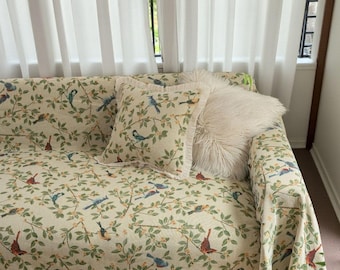 May include: A sofa with a cream-colored bird and floral patterned cover and matching throw pillows. The throw pillows include a square pillow with a bird and floral pattern and a fluffy white pillow. The sofa is near a window with white curtains.