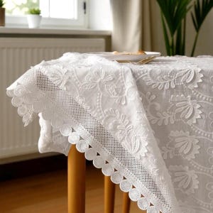 Op de afbeelding: Wit geborduurd tafelkleed met een bloemmotief. Het tafelkleed heeft een geschulpte rand met een decoratieve rand. De tafel is van hout en er staat een bord met eten op.