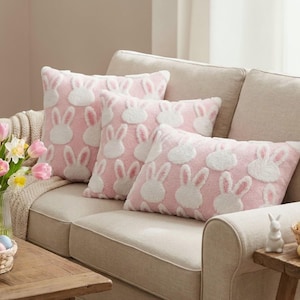 Puede incluir: Cojines decorativos rosas con diseños de conejos blancos. Los cojines están sobre un sofá beige. Las caras de los conejos están bordadas con pelo blanco y orejas internas rosas. Los cojines son rectangulares y cuadrados.
