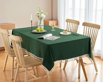 May include: A dark green rectangular tablecloth with a white fringe trim, set on a wooden table. The table is surrounded by four wooden chairs. A vase of flowers, a candle, and a plate of fruit are on the table.